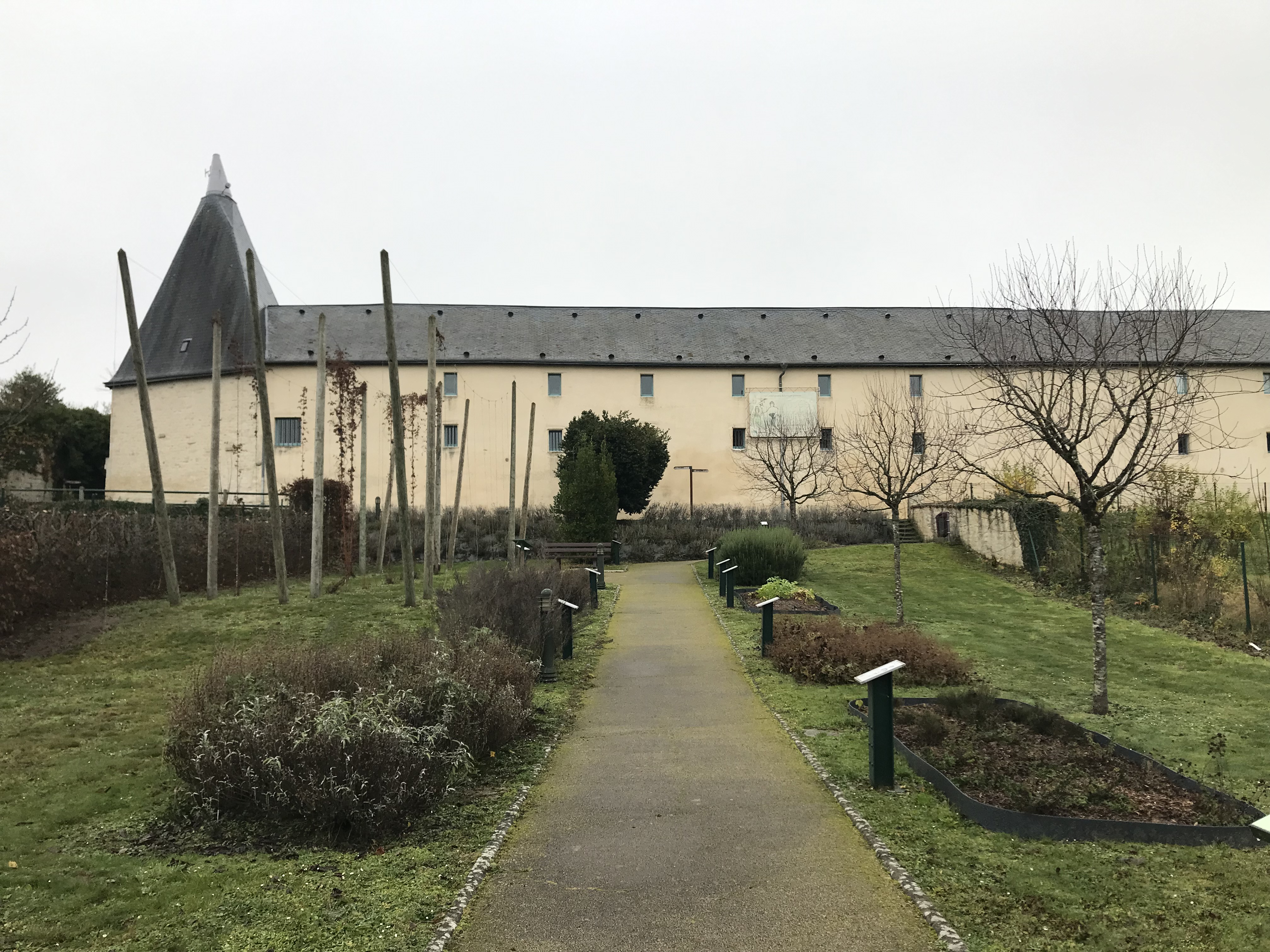 grand corps de bâtiment longitudinal en arrière-paln avec une tour à l'extémité gauche. Devant, le jardin avec les plantes utilisées dans la fabrication de la bière