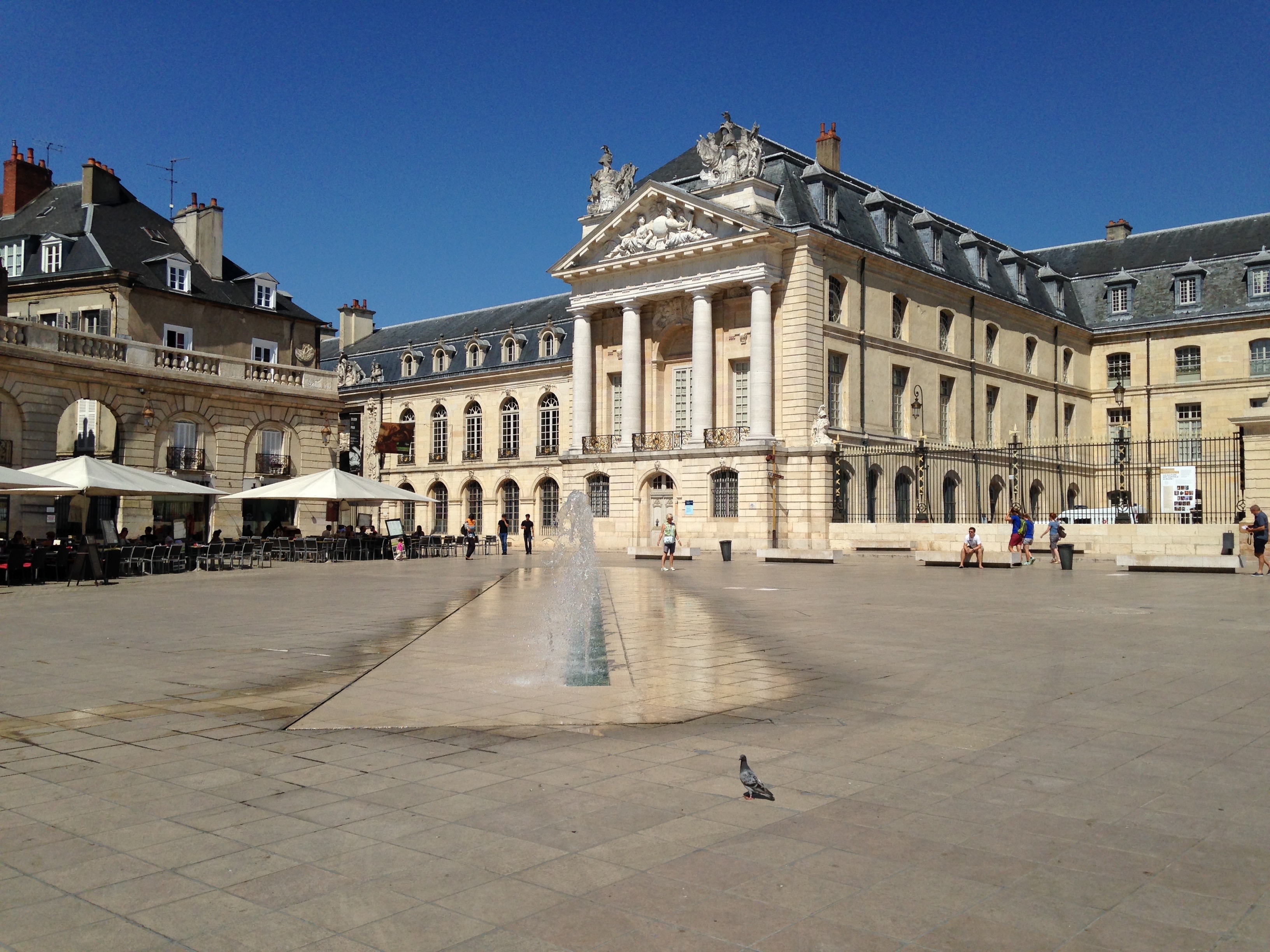 palais néo-classique avec grande place pavée devant, vue de biais, avec rangée de jets d'eau au premier plan